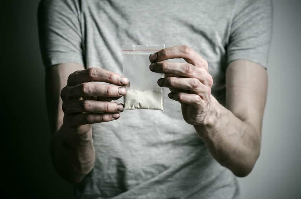 A man in a gray shirt holds a package of cocaine and questions his addiction.