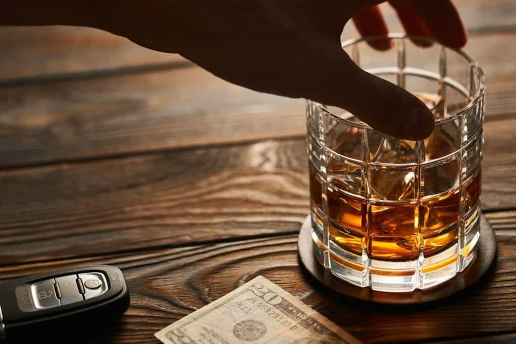 Man’s hand sets down glass of whisky next to car keys implying he intends to drive after drinking.